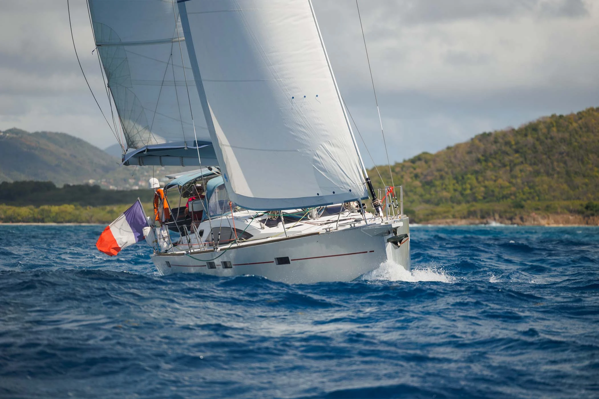 Voilier de grande croisière Allures avec couple à bord, en navigation autonome le long d’une côte tropicale