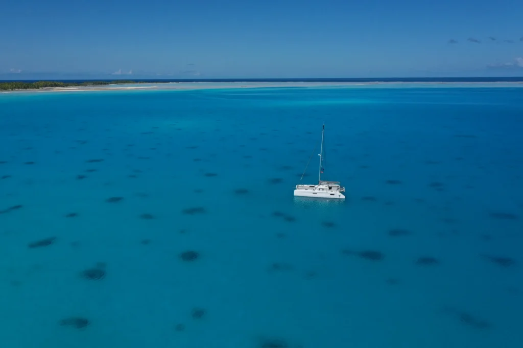 Photo aérienne d’un catamaran Outremer blanc nommé Ningyo, seul au mouillage dans un vaste lagon turquoise, avec des taches sombres de corail visibles sous la surface et un rivage de sable bordé de cocotiers à l’horizon.