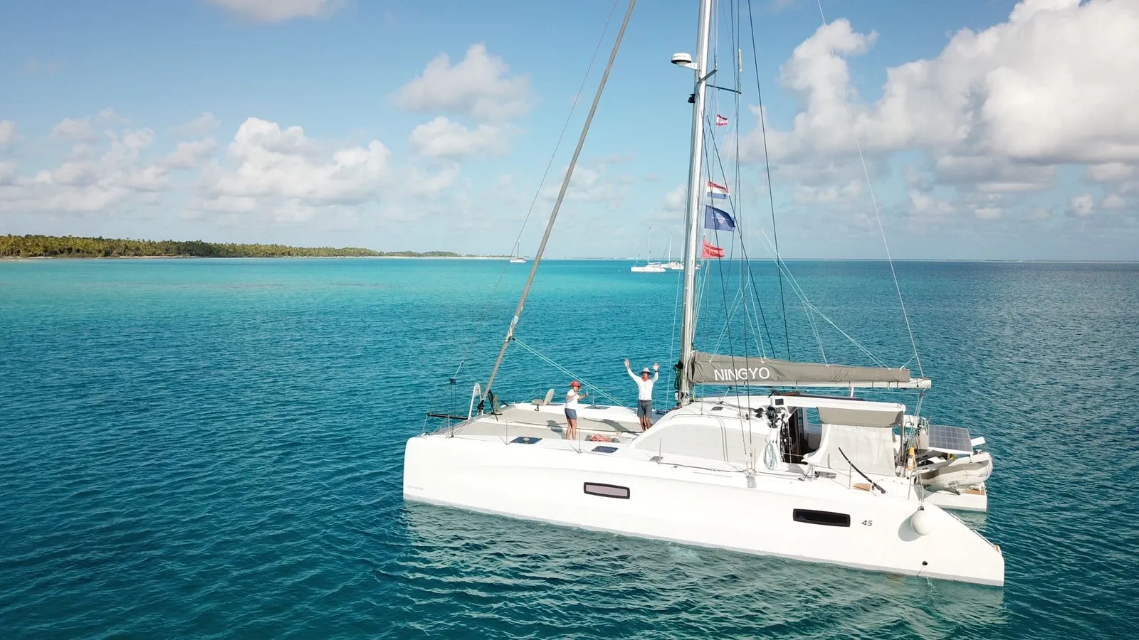 Ningyo au cœur d’un lagon turquoise des Tuamotu Vue aérienne du catamaran Outremer Ningyo, ancré dans les eaux turquoise d’un lagon polynésien. À bord, l'équipage saluent depuis le pont, entourés par un environnement marin paisible et préservé.